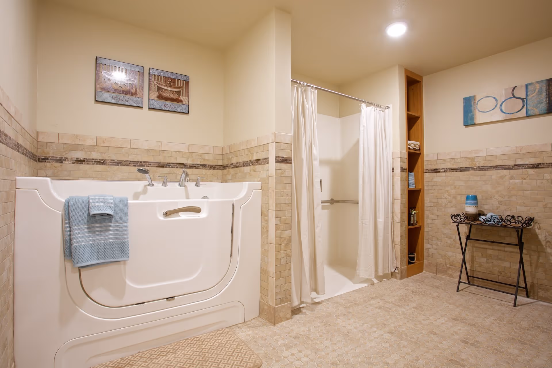 A spacious bathroom featuring a walk-in bathtub with a blue towel hanging on the side and a walk-in shower with white curtains. The walls are tiled halfway up with beige and brown tiles, and there are two framed pictures above the bathtub. A small black metal table with rolled blue towels and a decorative vase is placed against the wall, and there is a built-in wooden shelving unit with towels and toiletries.