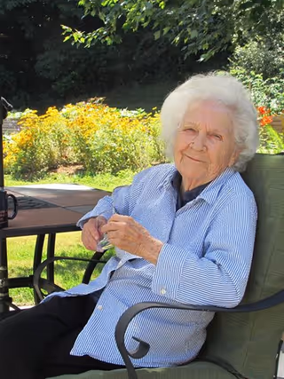 An elderly woman with white hair sitting outdoors on a green cushioned chair. She is wearing a blue and white striped shirt and holding a pair of glasses. Behind her, there is a garden with yellow flowers and green trees.