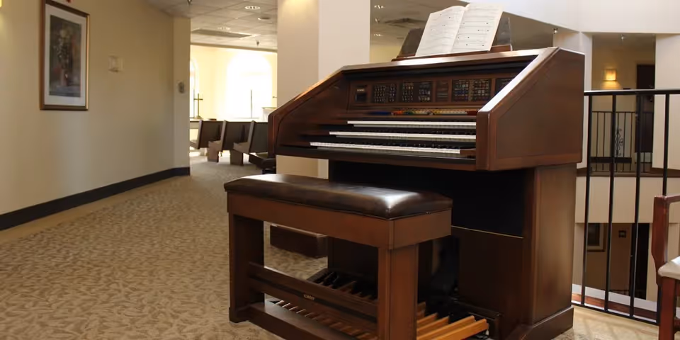 A wooden electronic organ with a padded bench is placed in a carpeted hallway of a nursing and rehabilitation center. The organ has sheet music on its stand. The hallway has beige walls, a framed picture on the left wall, and seating visible in the background near large windows letting in natural light.