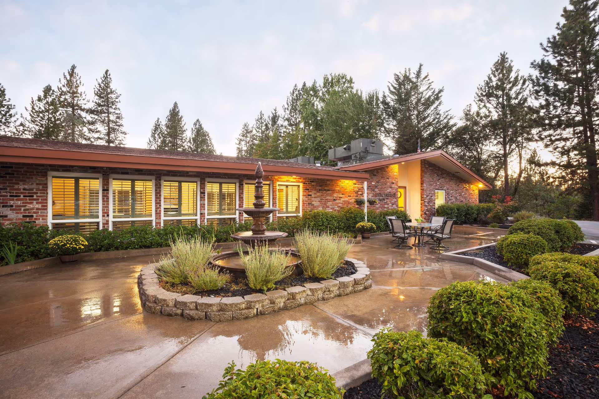 Exterior view of The Pines at Placerville Healthcare Center during dusk, showing a brick building with large windows illuminated from inside. In front of the building is a circular stone planter with a tiered fountain surrounded by greenery and bushes. There is a wet concrete patio area with outdoor seating including a table and chairs. Tall trees and shrubs surround the area.