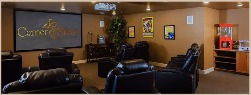 A cozy movie room with multiple black leather recliners arranged in rows facing a large screen displaying the CornerStone Assisted Living Community logo. The room has warm brown walls, a popcorn machine on a wooden cabinet, framed posters on the wall, and a ceiling-mounted projector.