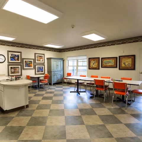 A bright dining room with checkered flooring, several tables and orange chairs arranged around them, a small kitchen island with a sink, framed artwork on the walls, and a large window letting in natural light.