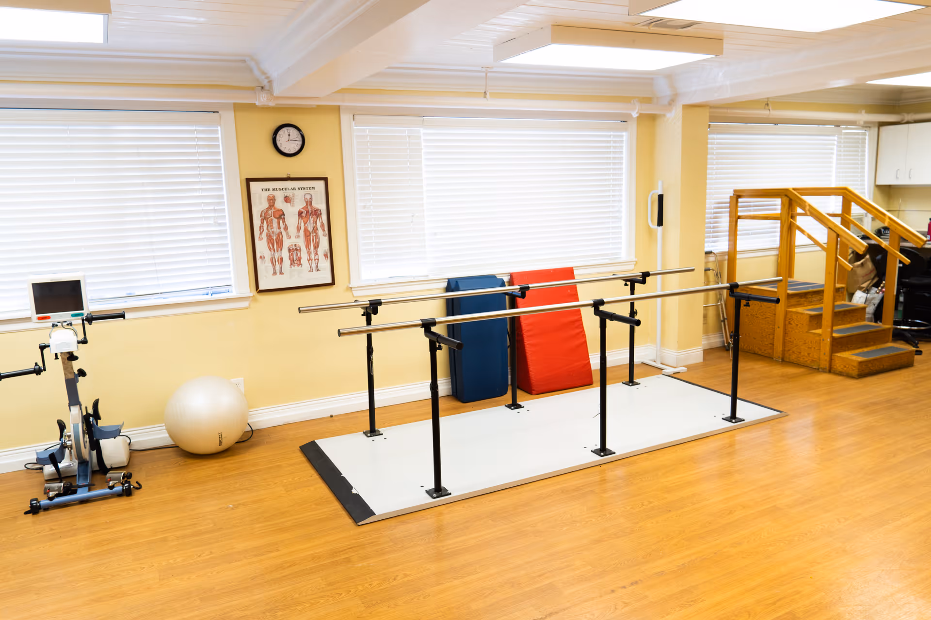 A rehabilitation room with parallel bars for walking exercises, a stationary exercise bike, a large exercise ball, padded mats leaning against the wall, and a small wooden staircase for physical therapy. The room has light yellow walls, large windows with white blinds, and a wooden floor.