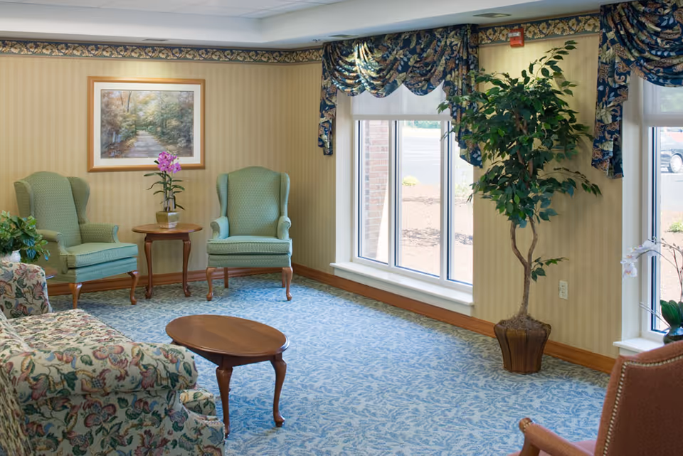 A cozy sitting area in a senior living facility with two green upholstered armchairs, a floral patterned sofa, a small wooden coffee table, and a round side table with a potted orchid. The room has large windows with floral valances, a framed nature painting on the wall, and a tall potted plant near the window.