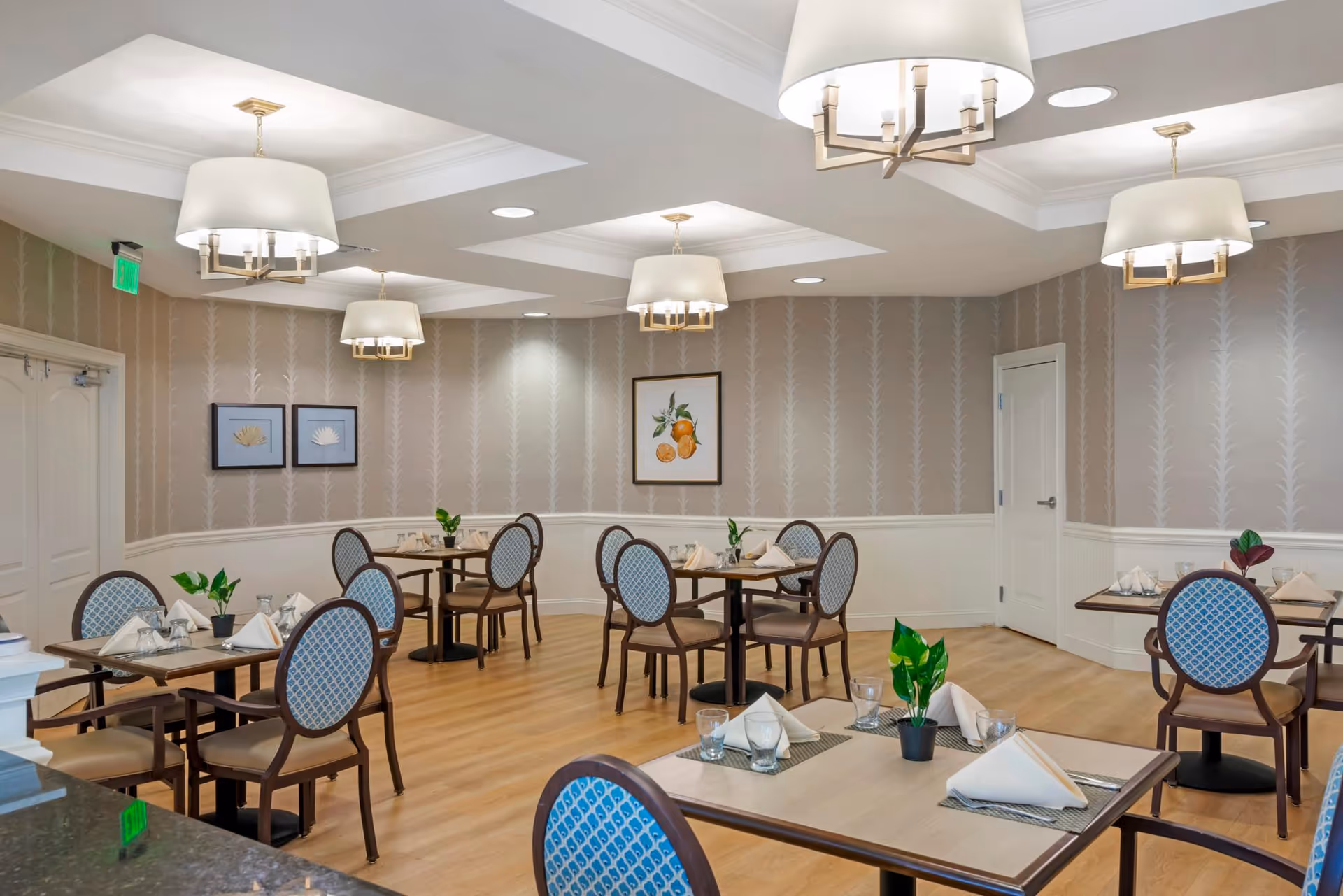 A dining room with several tables and chairs arranged neatly. Each table has a small plant, folded napkins, and glassware. The room features light wood flooring, patterned wallpaper, and multiple ceiling light fixtures with white lampshades. There are framed pictures on the walls and two closed doors visible.