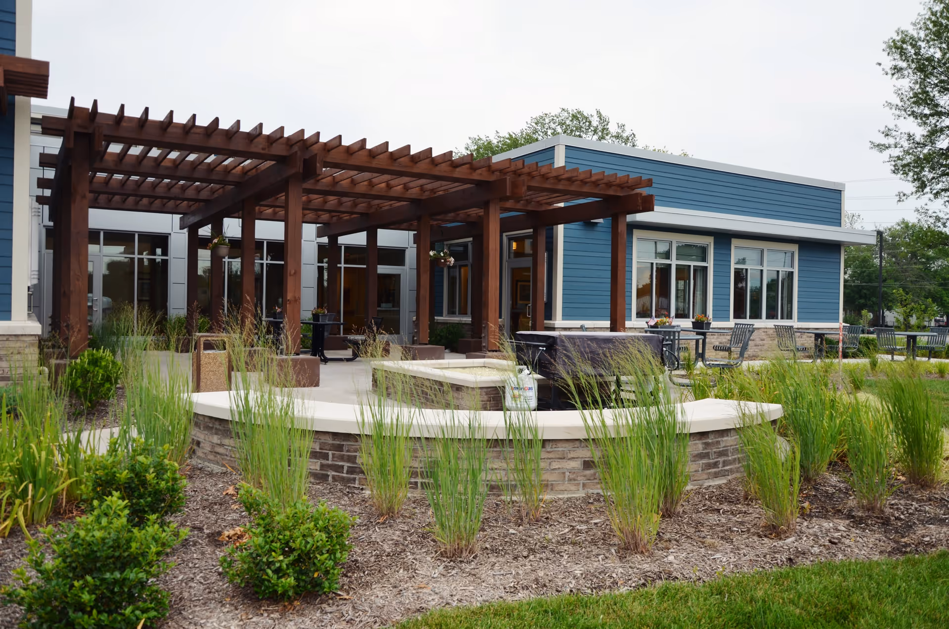 Outdoor patio area at Arlington Place Health Campus featuring a wooden pergola, seating areas with tables and chairs, surrounded by landscaped greenery and plants.
