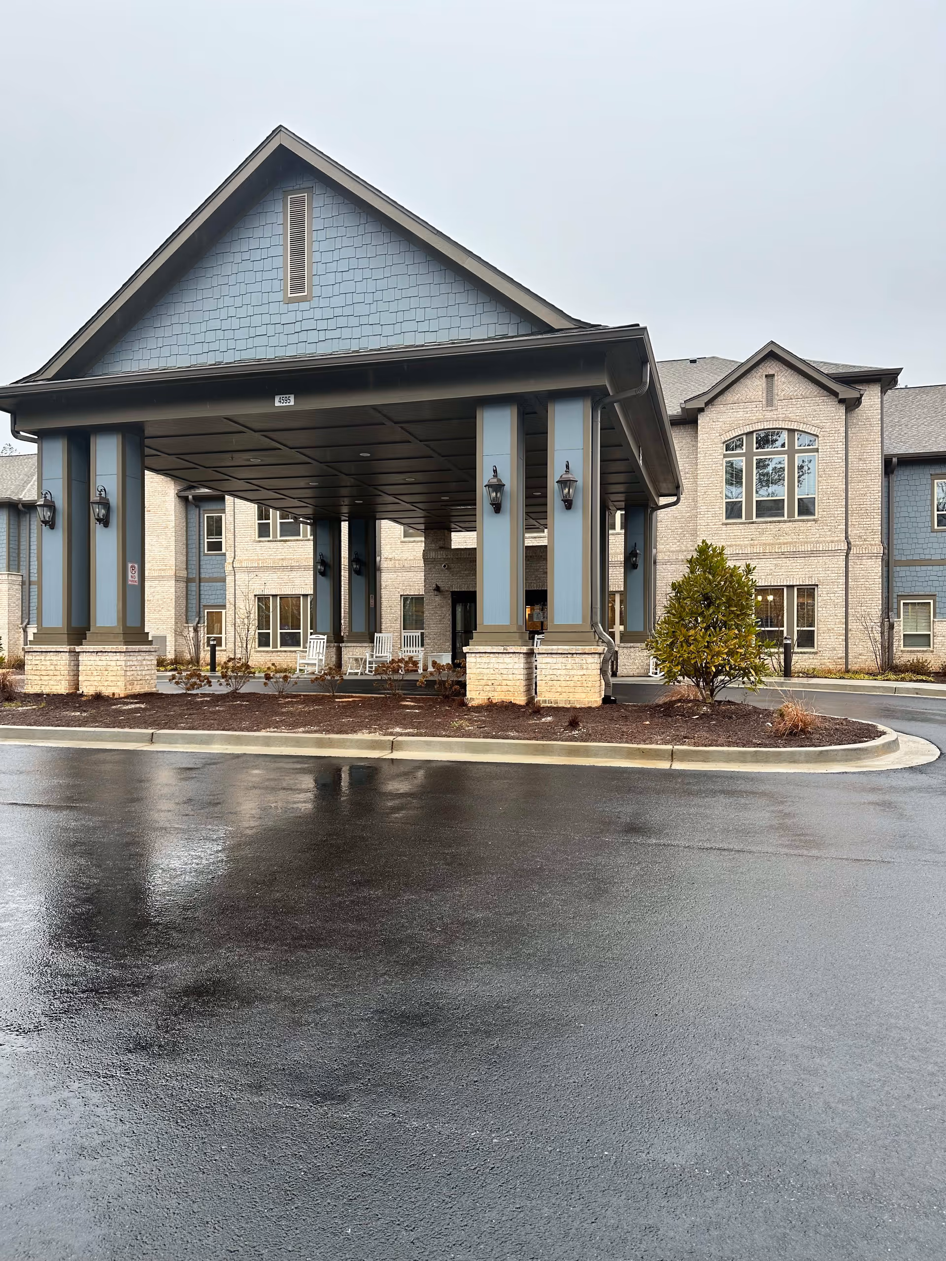 Front exterior view of a senior living facility with a covered entrance supported by columns. The building has light brick walls with blue paneling and multiple windows. There are rocking chairs under the covered entrance and a small landscaped area with a shrub in front. The ground is wet, indicating recent rain.