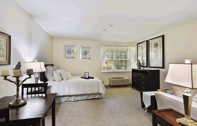 A bright and spacious senior living bedroom featuring a neatly made bed with multiple pillows, a window with sheer curtains allowing natural light, a dark wood dresser with decorative items, framed botanical artwork on the walls, a small table with a lamp and candles, and a comfortable armchair with a side table and lamp.