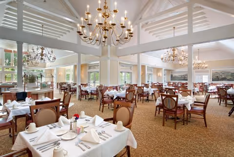 A spacious and elegant dining room with multiple tables covered in white tablecloths, set with plates, cups, silverware, and folded napkins. The room features high vaulted ceilings with exposed beams and several large chandeliers providing warm lighting. Large windows allow natural light to fill the space, and framed artwork is visible on the walls.