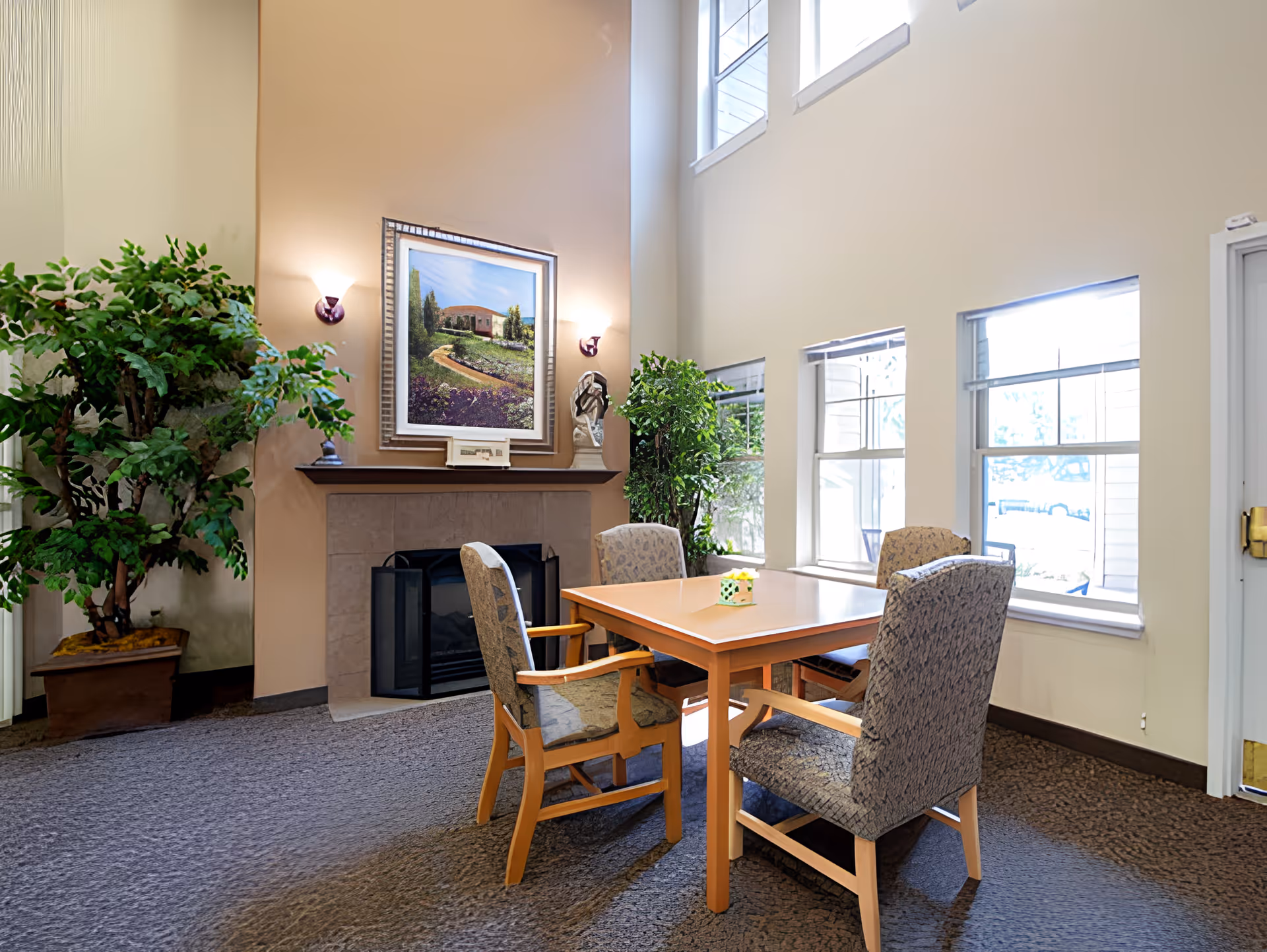 A cozy sitting area with a wooden table and four upholstered chairs arranged around it. The room features a fireplace with a framed painting above it, two wall sconces, and large windows letting in natural light. There are also two large potted plants adding greenery to the space.