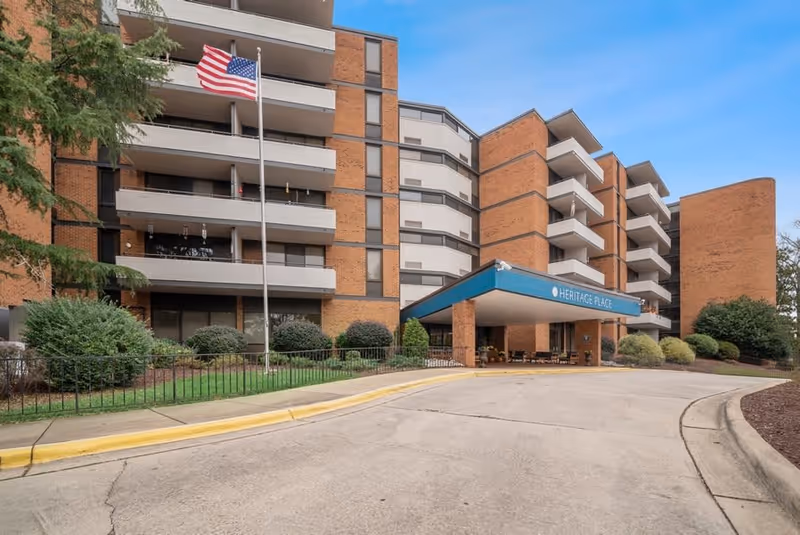 Exterior view of a multi-story assisted living facility building with brick and white paneling. The entrance has a blue canopy with the text 'HERITAGE PLACE'. An American flag is flying on a flagpole in front of the building, and there are bushes and trees around the entrance area.