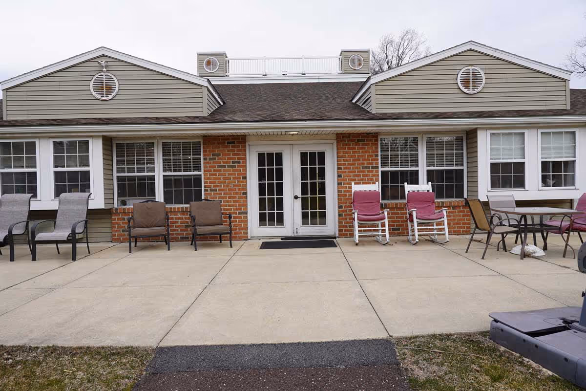 Exterior view of a single-story building with a brick and siding facade, featuring a central double glass door entrance flanked by multiple windows. Several outdoor chairs and a table are arranged on the concrete patio in front of the building.