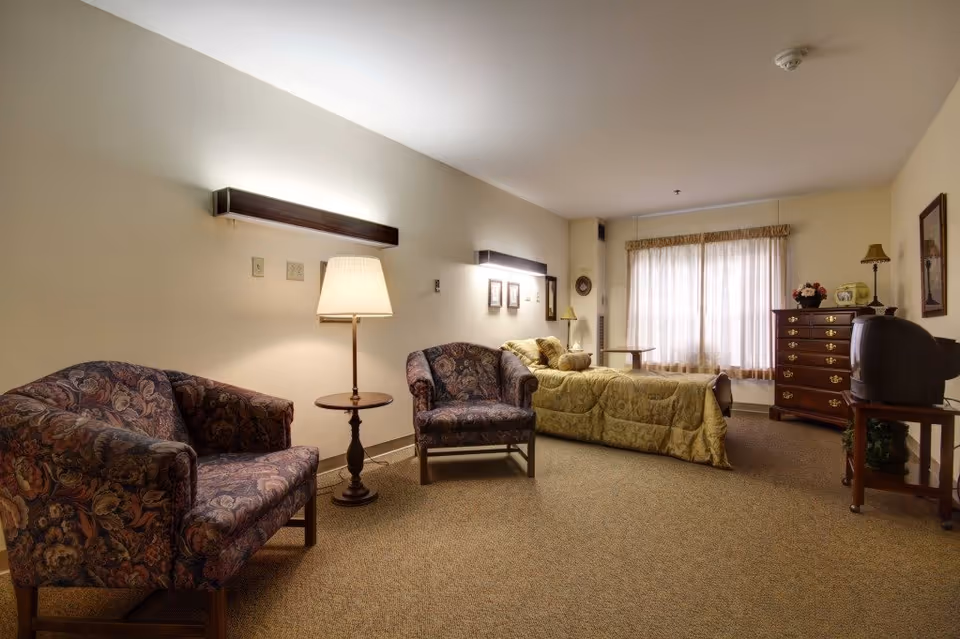 A cozy assisted living bedroom featuring a single bed with a gold bedspread, two patterned armchairs, a wooden side table with a lamp, a wooden dresser with a lamp and decorative items, a TV on a wooden stand, and a window with sheer curtains allowing natural light to fill the room.