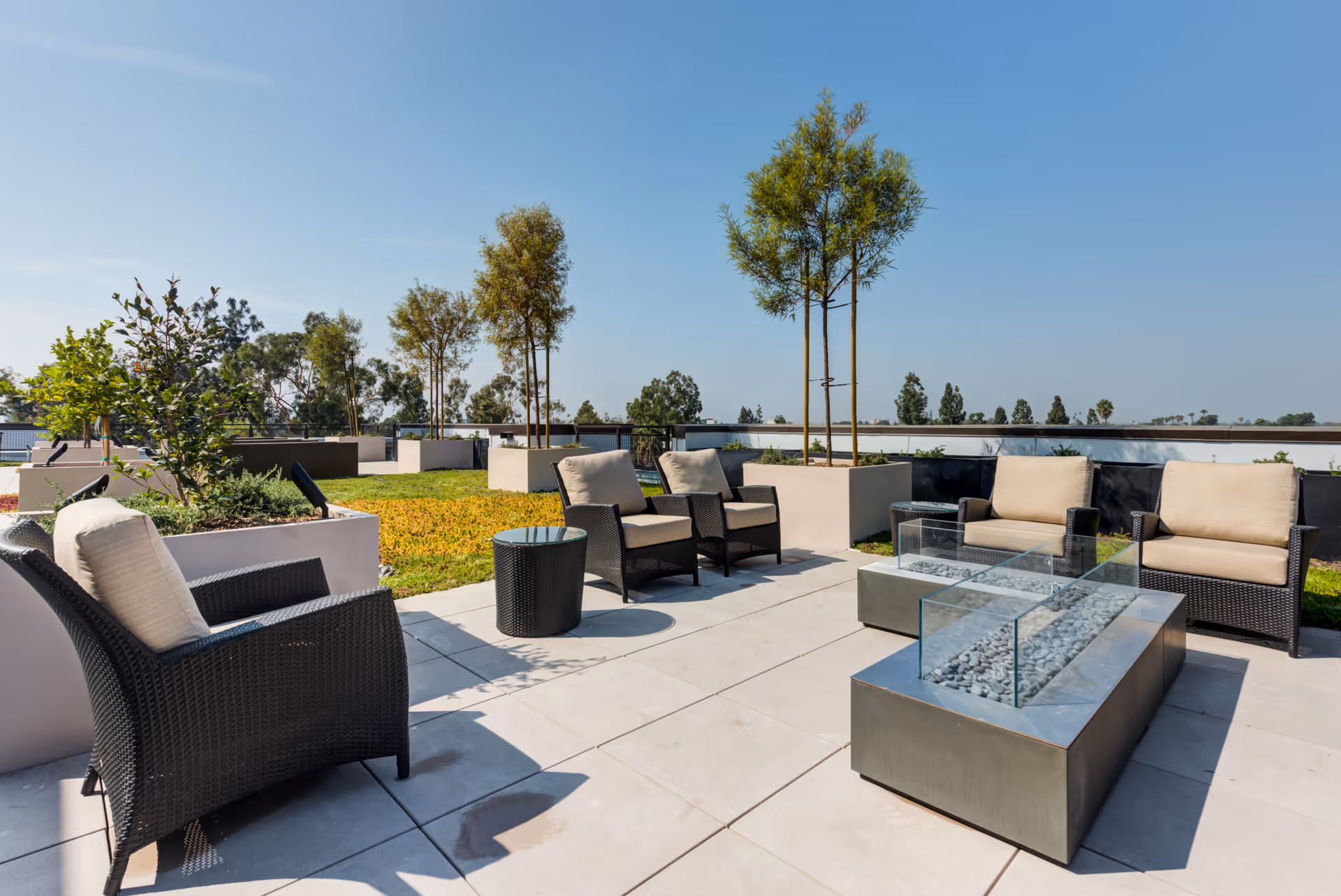 Rooftop patio with wicker seating, glass-enclosed fire pits, planters and small trees under a clear blue sky.
