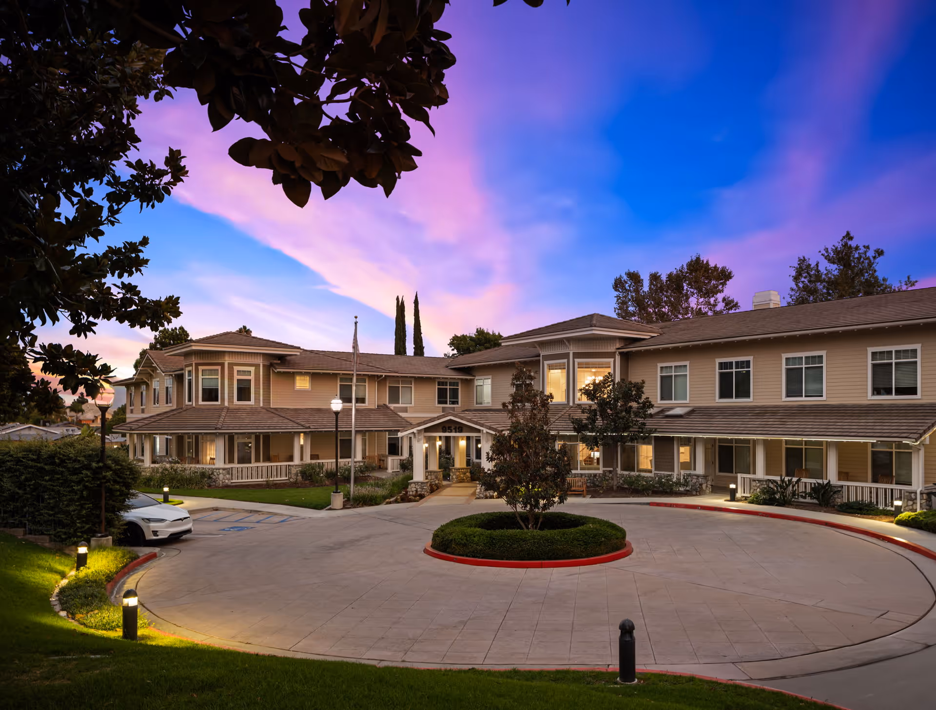 Exterior view of a two-story senior living facility building at dusk with a circular driveway and landscaped greenery, under a vibrant purple and blue sky.
