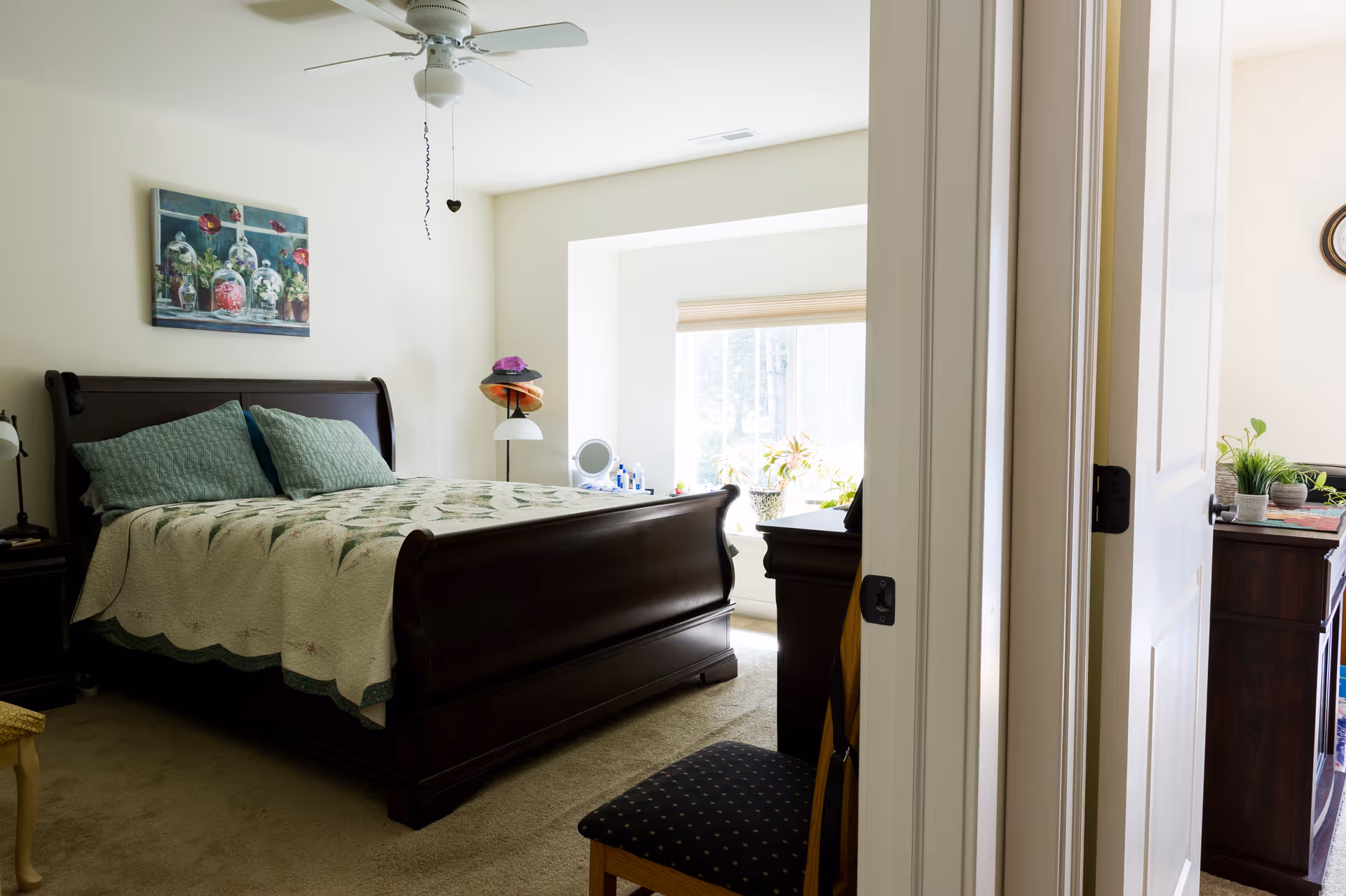 Sunlit bedroom with a dark wooden sleigh bed covered by a quilt, a window seat with plants, dressers and an open door in the foreground.