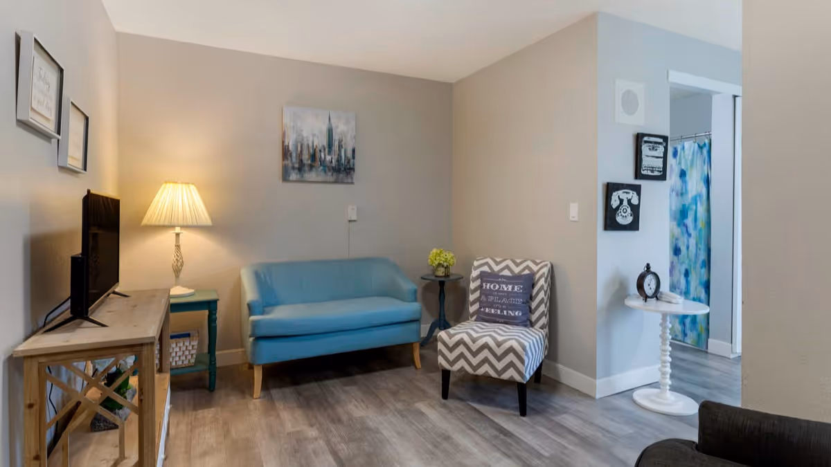 A cozy living area with a light blue loveseat, a chevron-patterned armchair with a decorative pillow, a wooden TV stand with a flat-screen TV, a side table with a lamp, and a small round table with a clock. The room has light gray walls and wood flooring. In the background, there is a glimpse of a bathroom with a colorful shower curtain.