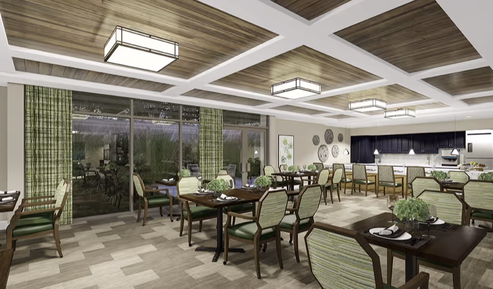 Spacious dining room with multiple wooden tables and green-upholstered chairs, large windows with green curtains, and a kitchen area in the back.