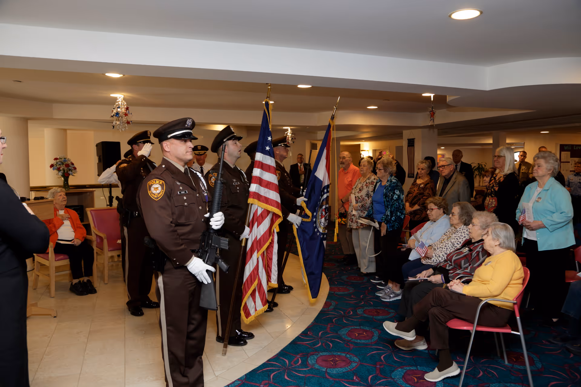 A group of uniformed police officers holding flags stand in formation inside a room, while a group of elderly people, some seated and some standing, watch respectfully. The room has a patterned carpet and overhead lighting.
