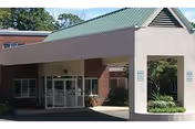 Entrance of Southeast Rehabilitation & Skilled Care Center showing a covered drop-off area with a green metal roof, glass double doors, and brick walls. There are signs on the pillars and some greenery around the building.