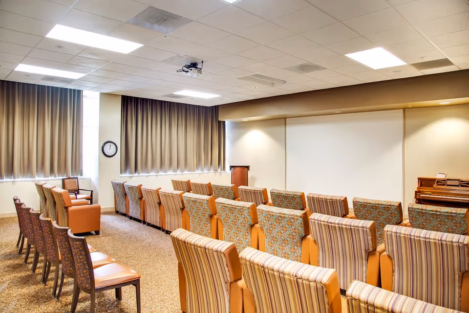 A well-lit room with rows of upholstered chairs facing a white projection screen. The room has beige walls, carpeted floor, and large windows with closed curtains. A clock is mounted on the wall near the windows, and a piano is visible in the corner.