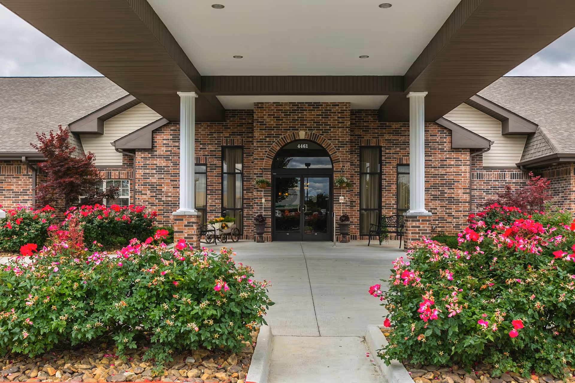 Entrance of a brick building with a covered porch supported by white columns. There are vibrant pink and red flowering bushes on either side of the concrete walkway leading to double glass doors. The building has a gabled roof and windows flanking the entrance.