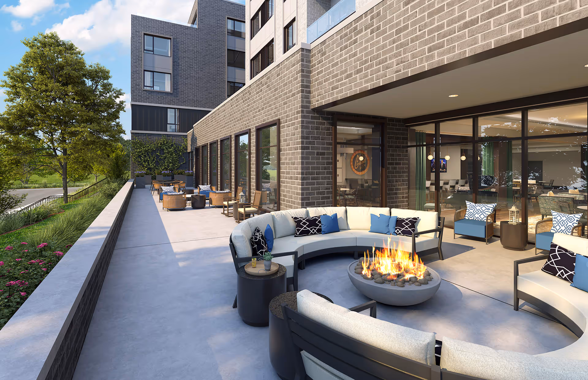 Outdoor patio area at The Sheridan at Oak Brook featuring a curved seating arrangement around a fire pit, additional seating with blue cushions, and tables. The patio is adjacent to a brick building with large windows showing an interior lounge area. There are trees and greenery along the edge of the patio.