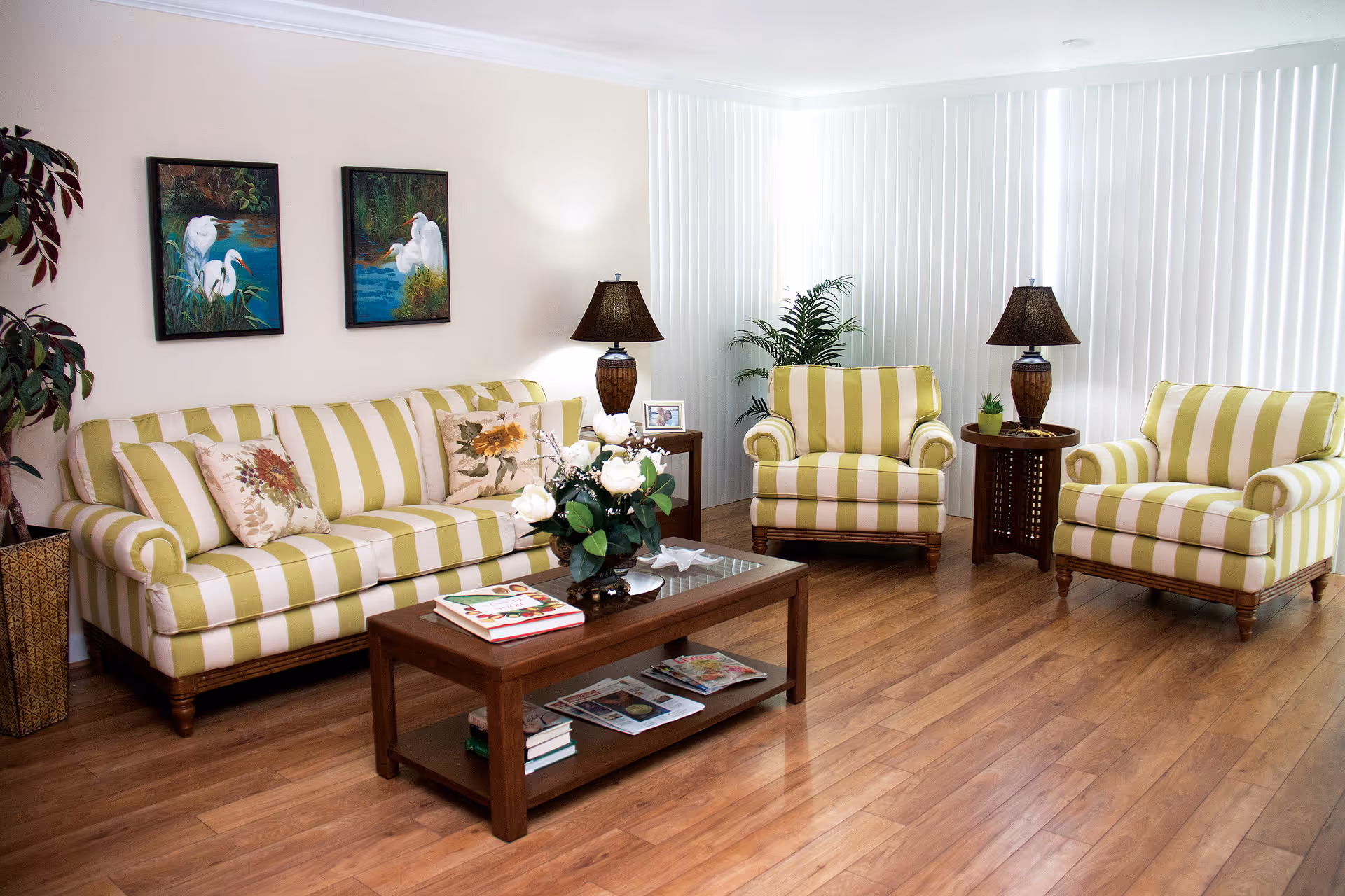 A bright living room with wooden flooring featuring a striped green and white sofa and two matching armchairs. There is a wooden coffee table with books and a flower arrangement on it. Two table lamps with dark shades are placed on side tables next to the armchairs. Two paintings of white birds hang on the wall above the sofa, and vertical blinds cover the windows in the background. A potted plant is placed in the corner.