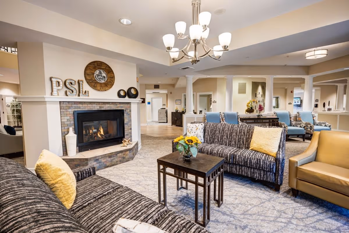 A cozy senior living facility common area with a lit fireplace surrounded by a stone hearth. The room features comfortable seating including patterned sofas with yellow and white pillows, a tan armchair, and blue cushioned chairs in the background. A wooden coffee table with a vase of sunflowers is centered in front of the sofas. The space is well-lit with ceiling lights and a chandelier, and decorated with wall art and decorative items on the fireplace mantel.