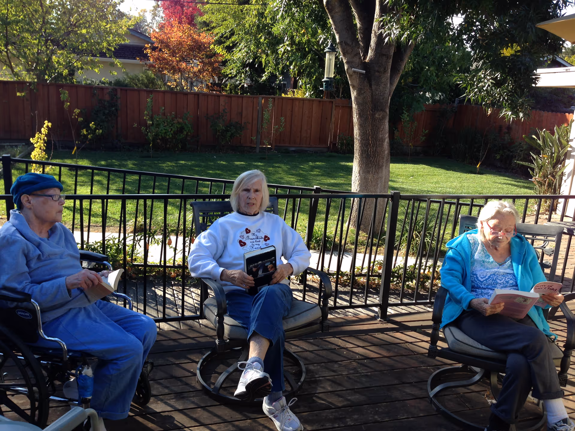 Three elderly women sitting outdoors on a wooden deck. One woman is in a wheelchair wearing a blue outfit and hat, holding a book. The second woman is sitting in a chair wearing a white sweatshirt and blue jeans, holding a book. The third woman is also seated, wearing a blue jacket and reading a book. Behind them is a fenced grassy yard with trees and plants.
