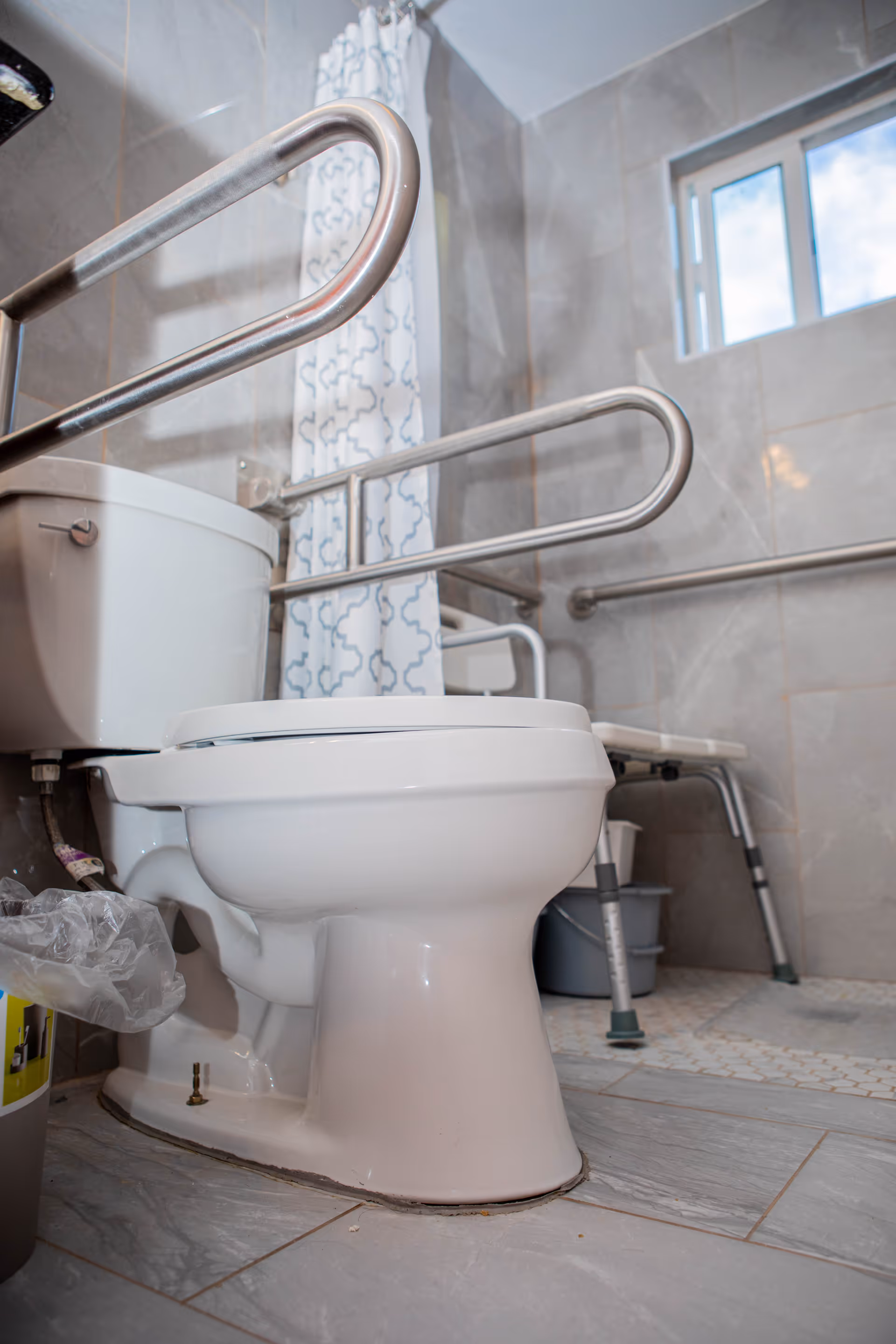 A bathroom with a white toilet equipped with metal safety grab bars on both sides. There is a shower area with a white and blue patterned shower curtain, a small window, and a shower chair with metal legs and green rubber tips. The walls and floor are tiled in gray tones.