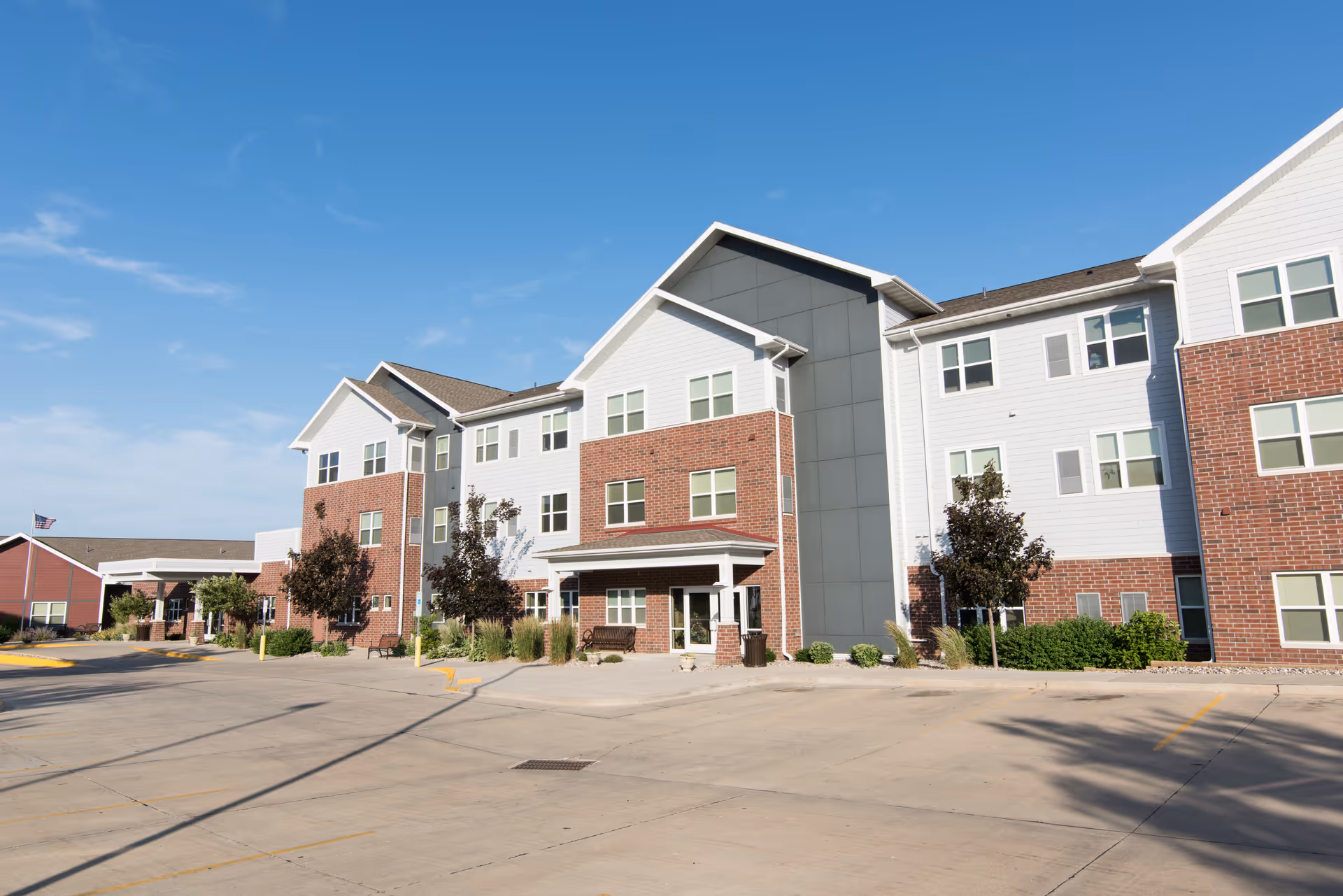 Front view of a three-story brick-and-siding senior living building with a large empty parking lot under a clear blue sky.