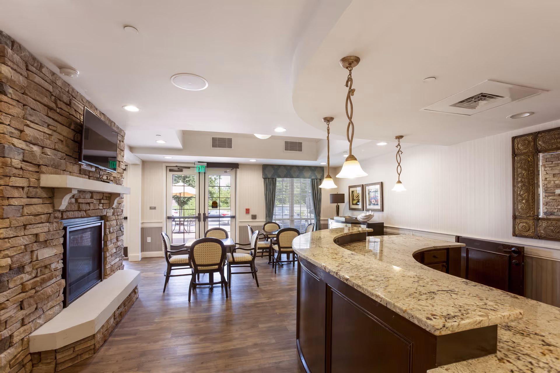 A cozy interior common area with a stone fireplace and mounted TV on the left, several round tables with chairs in the center near large windows and glass doors, and a curved granite countertop bar with pendant lights hanging above on the right.
