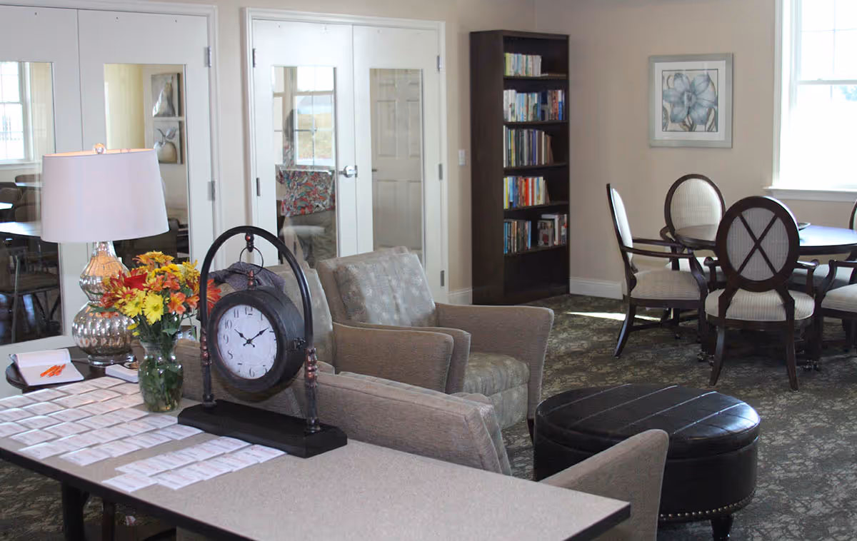 A cozy common area in a senior living facility featuring a table with a large clock, a vase of flowers, and name tags. There are two armchairs, a round table with four chairs, a bookshelf filled with books, and a window letting in natural light.