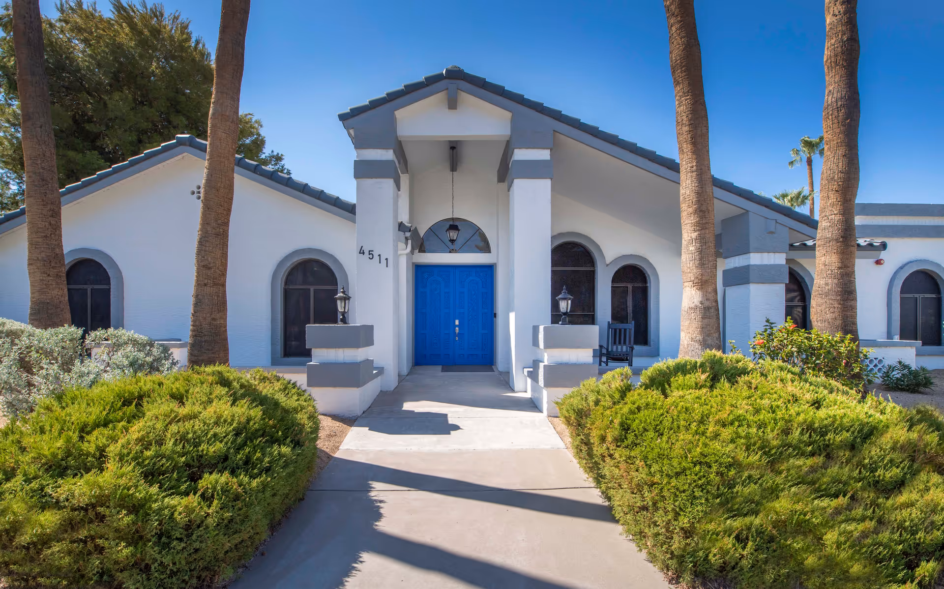 Front exterior view of a white building with gray trim and a blue double door entrance, flanked by two columns and lantern-style lights. The building is surrounded by green bushes and tall palm trees under a clear blue sky.
