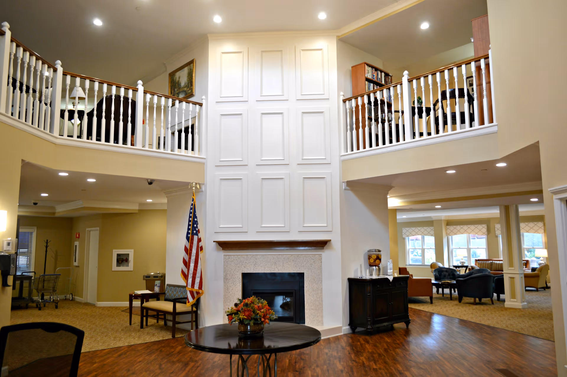 Interior view of a senior living facility common area with a high ceiling and white paneled wall above a fireplace. There is a round table with a flower arrangement in the center, an American flag to the left, and seating areas with chairs and sofas visible in the background. The upper level has white railings and bookshelves.