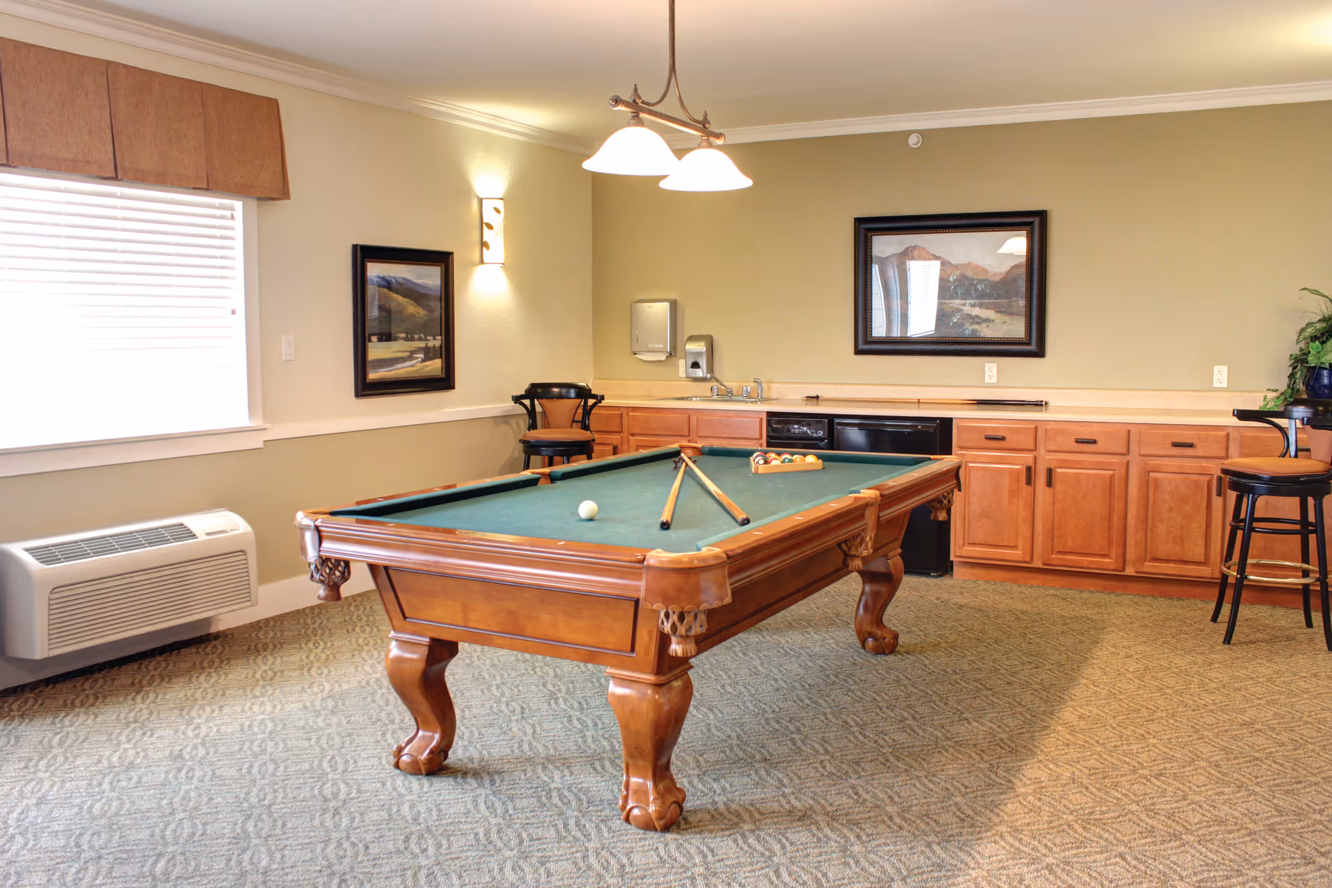 A recreational room with a wooden pool table in the center, two pool cues resting on the table, and a rack of billiard balls. The room has green carpet, light green walls, and wooden cabinets along the back wall with a sink and dishwasher. There are two framed landscape paintings on the walls, a window with blinds on the left, and two bar stools near the cabinets.