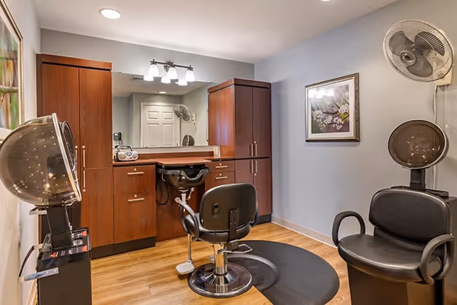 Interior of a hair salon room with wooden cabinets, a large mirror, two black salon chairs, a hair dryer, and a wall-mounted fan. The room has light-colored walls and wooden flooring, with a framed picture hanging on the wall.