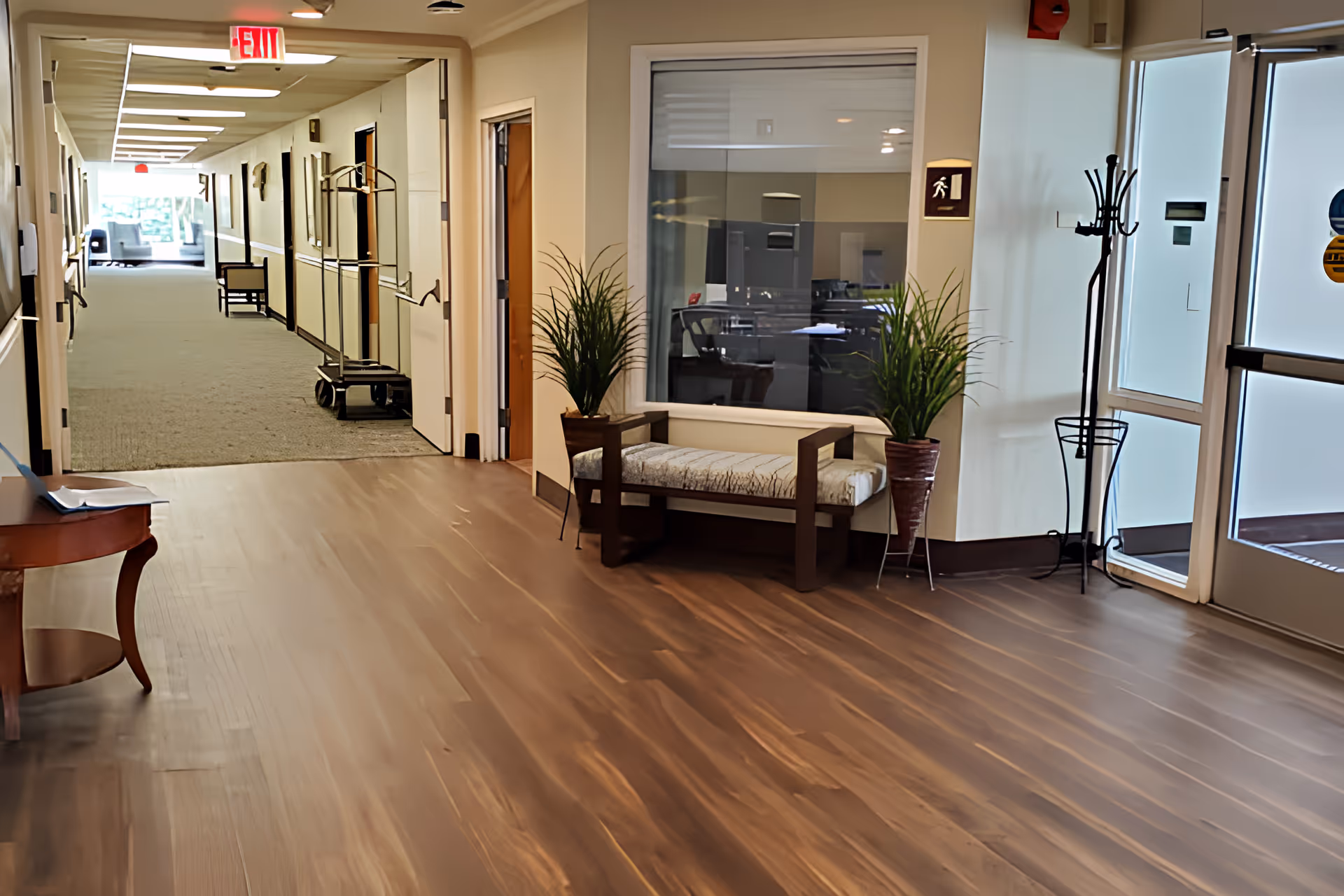 Interior view of a senior living facility hallway with wooden flooring near the entrance. There is a cushioned bench with two potted plants on either side, a coat rack, and a small wooden table. The hallway extends into a carpeted corridor with chairs and a luggage cart visible. An exit sign is illuminated above the corridor entrance.