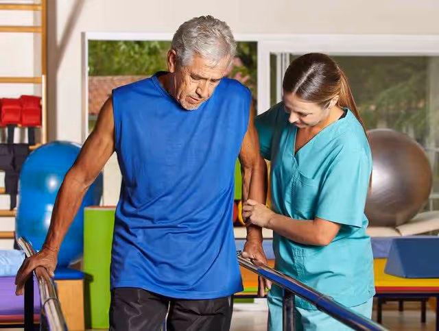 An elderly man in a blue sleeveless shirt is receiving physical therapy assistance from a female therapist wearing teal scrubs. They are in a rehabilitation or therapy room with exercise equipment such as large exercise balls and padded mats in the background.