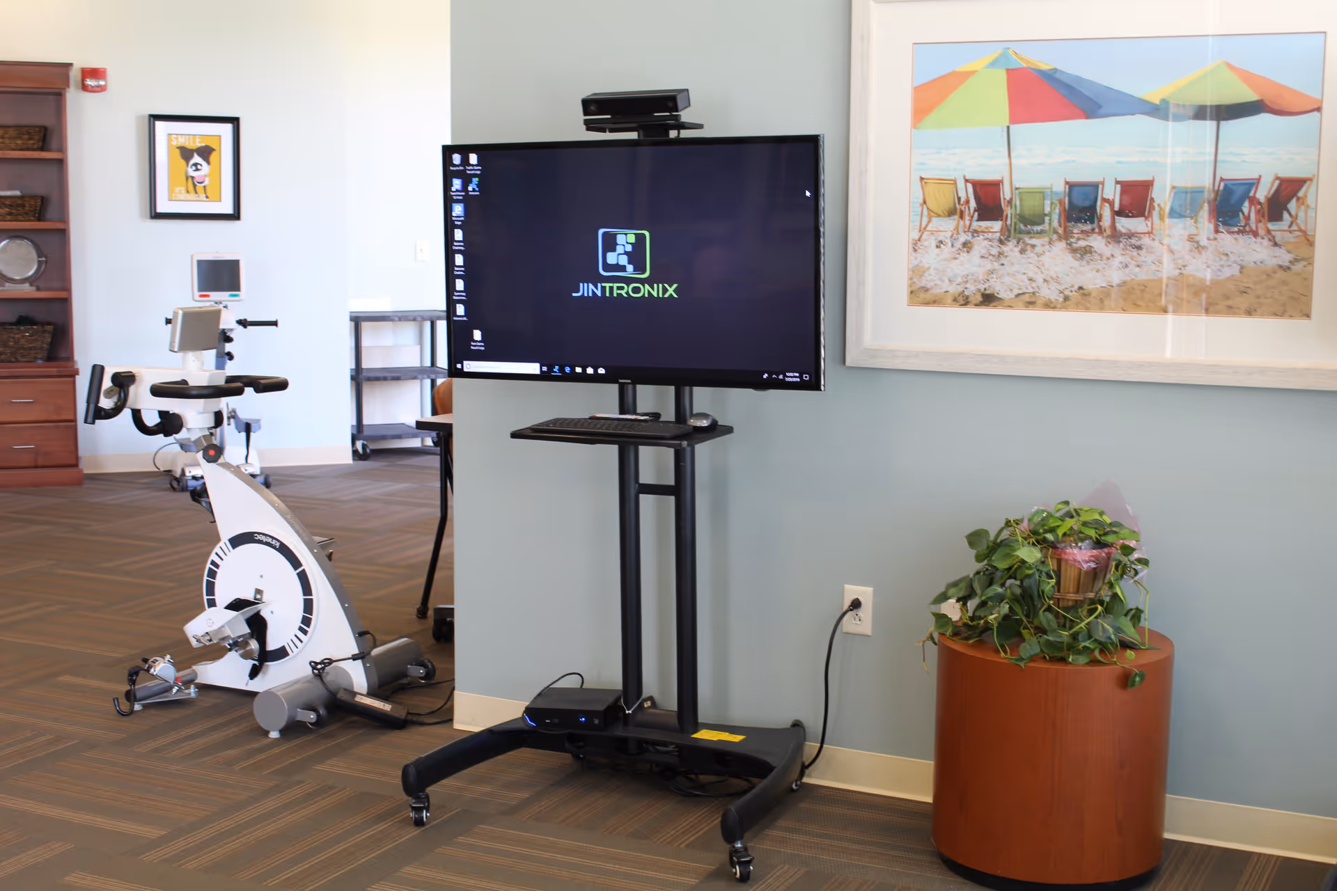 An interior room with a stationary exercise bike, a large monitor on a stand displaying the Jintronix logo, a framed picture of beach chairs and umbrellas on the wall, a small plant on a round wooden table, and a wooden bookshelf with baskets and a framed picture on the wall.