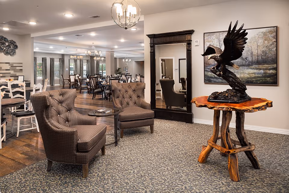 A senior living facility common area featuring two brown upholstered armchairs with a small round table between them, a wooden table with a large eagle sculpture on top, a tall ornate mirror, and a dining area with multiple tables and chairs in the background under chandeliers.