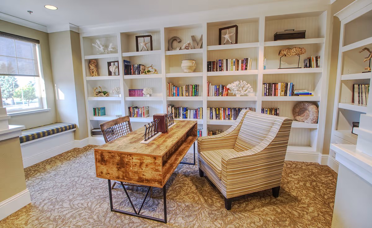 A cozy reading nook in Carmel Village Memory Care featuring a wooden desk with two chairs, one striped armchair and one wooden chair, set on a patterned carpet. Behind the desk is a large white built-in bookshelf filled with books and decorative items like coral, starfish, and shells. A window with a roller shade allows natural light into the room, and there is a cushioned window seat beneath it.