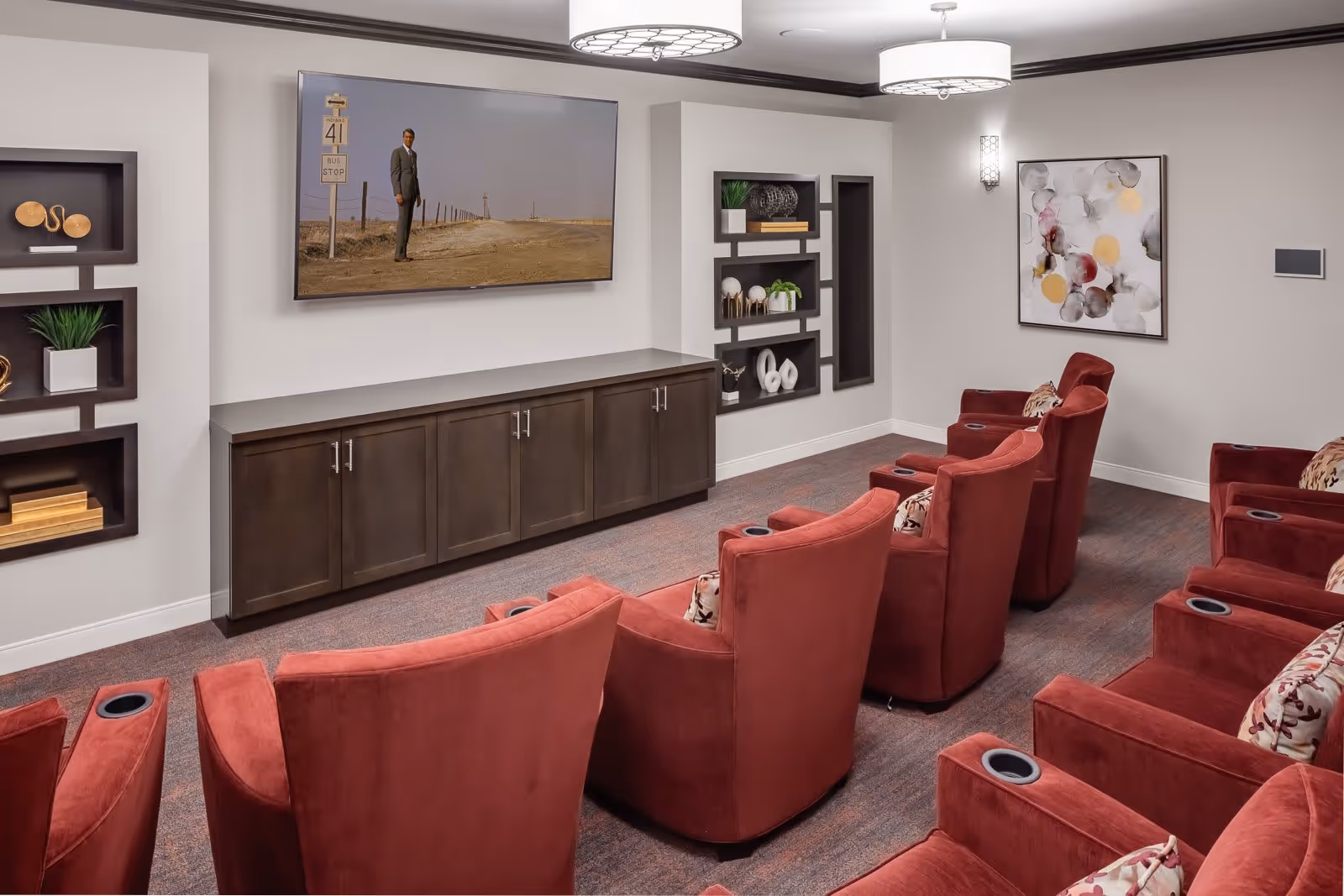 A cozy media room with two rows of red upholstered recliner chairs, each with built-in cup holders. A large flat-screen TV is mounted on the wall above a dark wooden cabinet. The walls are decorated with built-in shelves holding decorative items and a colorful abstract painting. The room has soft lighting from ceiling fixtures and a wall sconce.