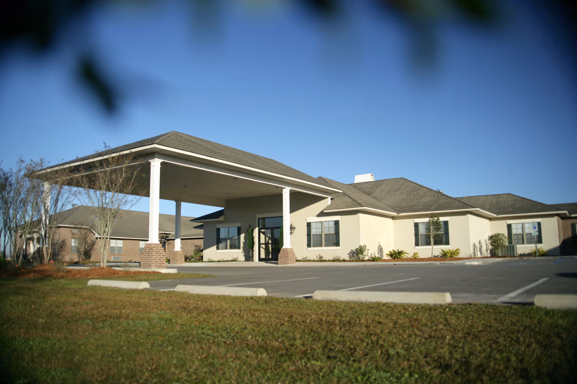 Exterior view of a single-story senior living facility building with a covered entrance supported by white columns. The building has beige walls, multiple windows, and a gray shingled roof. There is a parking lot in front with marked parking spaces and some landscaping with grass and small trees.