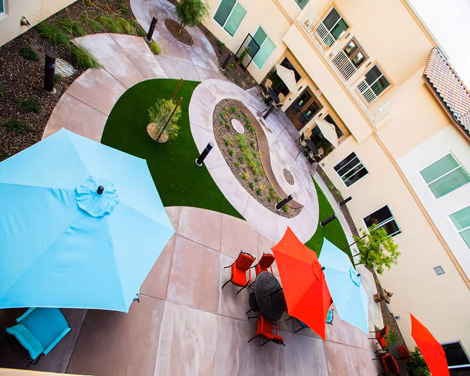 Outdoor courtyard area with curved walkways, landscaped garden beds, and several tables with colorful umbrellas in blue and red. The courtyard is surrounded by a beige building with multiple windows and doors, some with balconies and patio umbrellas.