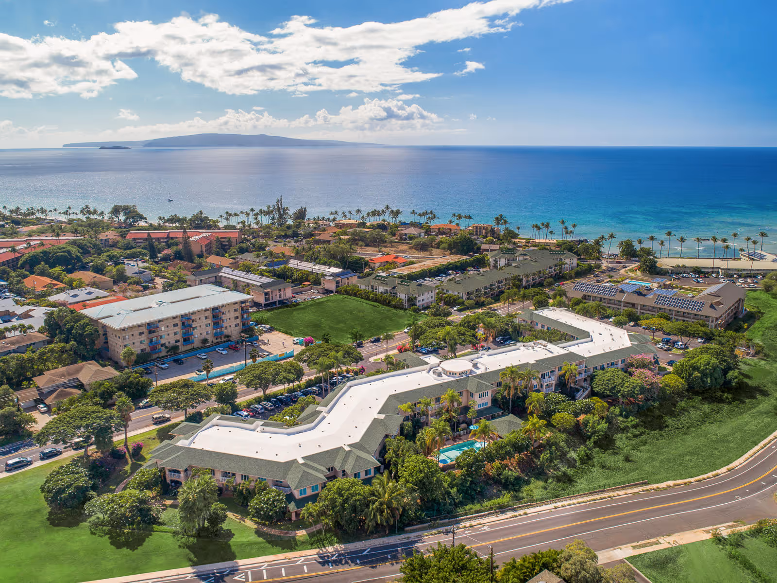 Aerial exterior view of Holiday Kalama Heights senior living complex with white-roofed buildings, pool, palm trees and the ocean beyond.