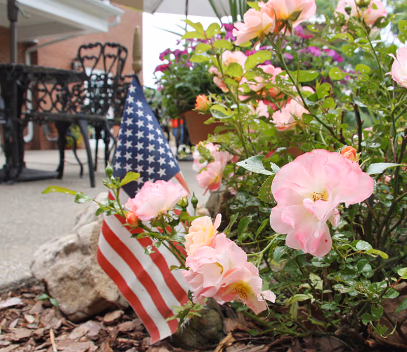 Close-up of pink flowers and an American flag planted in a garden bed with mulch and rocks, with outdoor metal chairs and a brick building visible in the background.