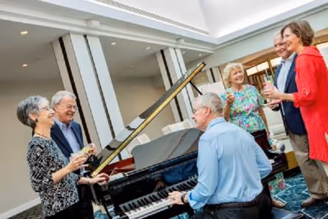 A group of six seniors socializing around a grand piano in a bright, spacious common area with large windows and modern decor. One man is playing the piano while others stand nearby holding drinks and smiling.