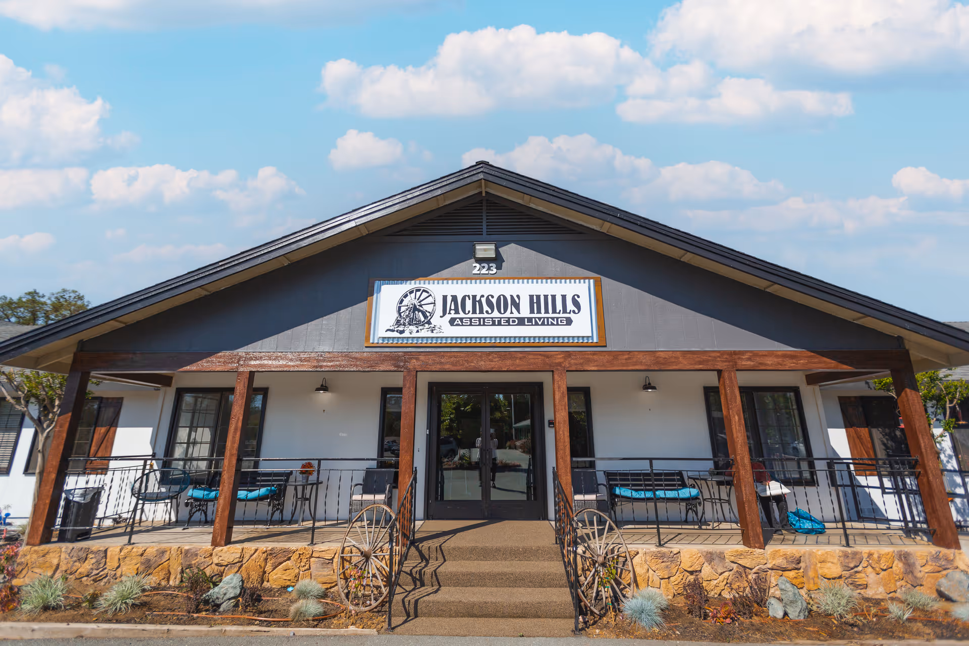 Front exterior view of Jackson Hills Assisted Living facility with a porch featuring wooden beams, seating areas with chairs and cushions, stone foundation, and a sign above the entrance displaying the facility name.